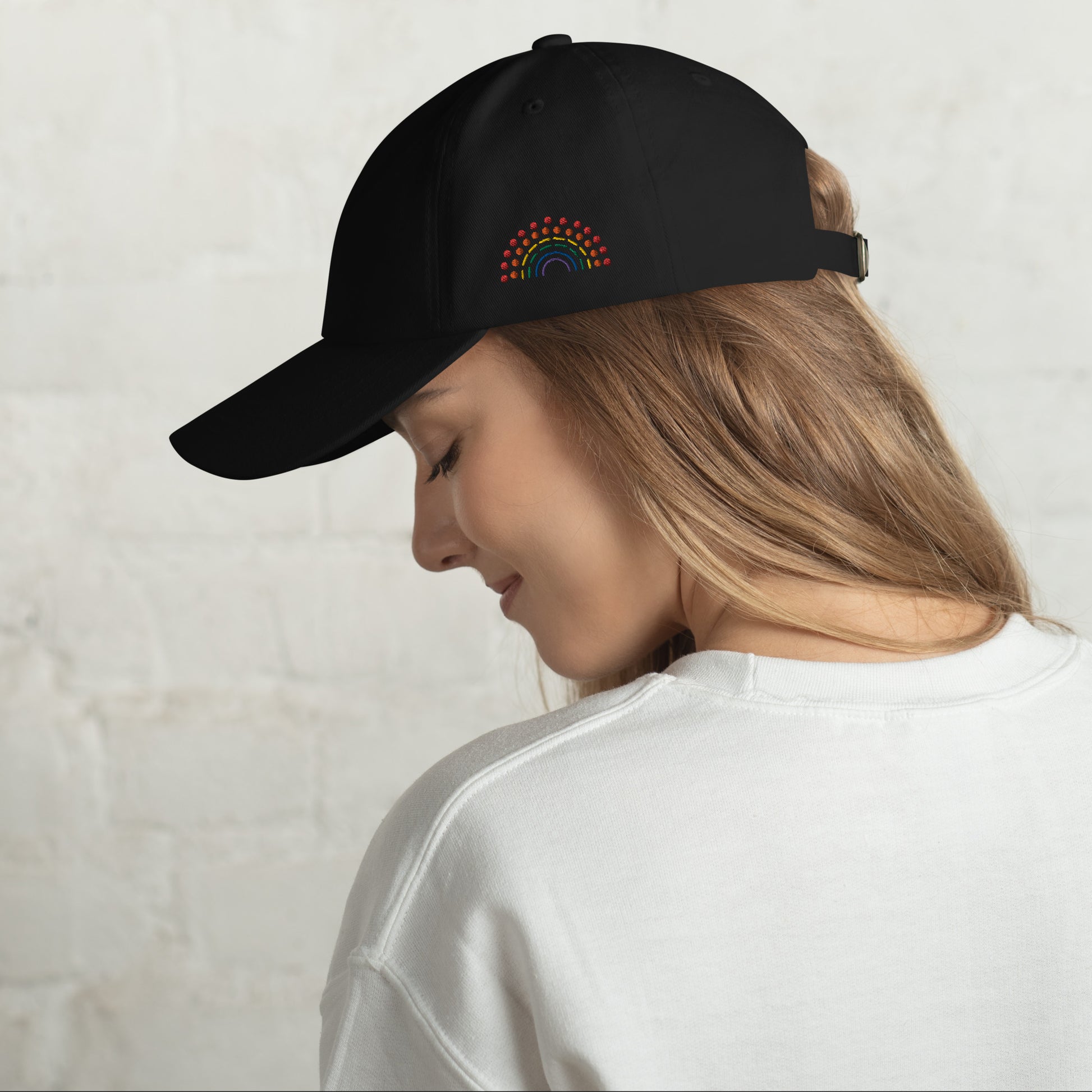 person wearing a black baseball dad hat with multicolored pride rainbow embroidered on the left of the hat