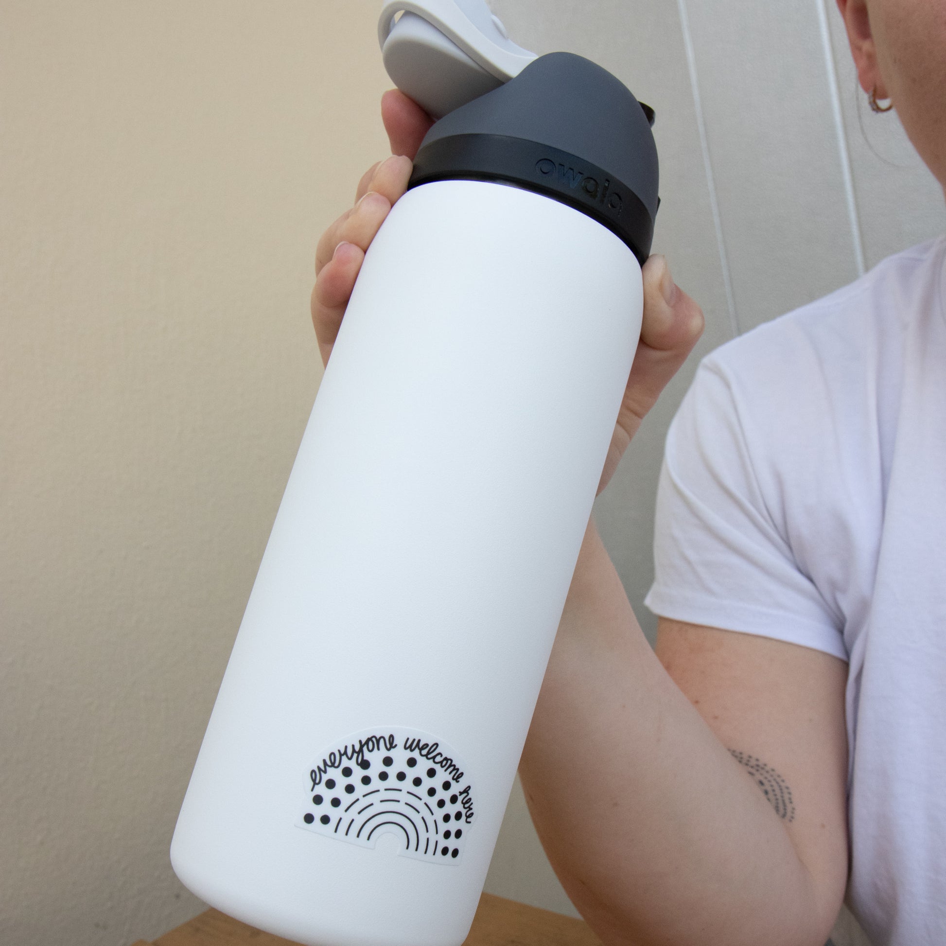 Person holding a water bottle with a black pride sticker with the phrase everyone welcome here