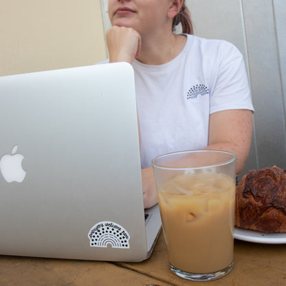 Person working on a laptop with a black pride sticker with the phrase everyone welcome here