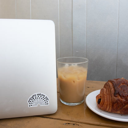 Laptop with a multicolored pride sticker with the phrase everyone welcome here amidst a coffee and croissant