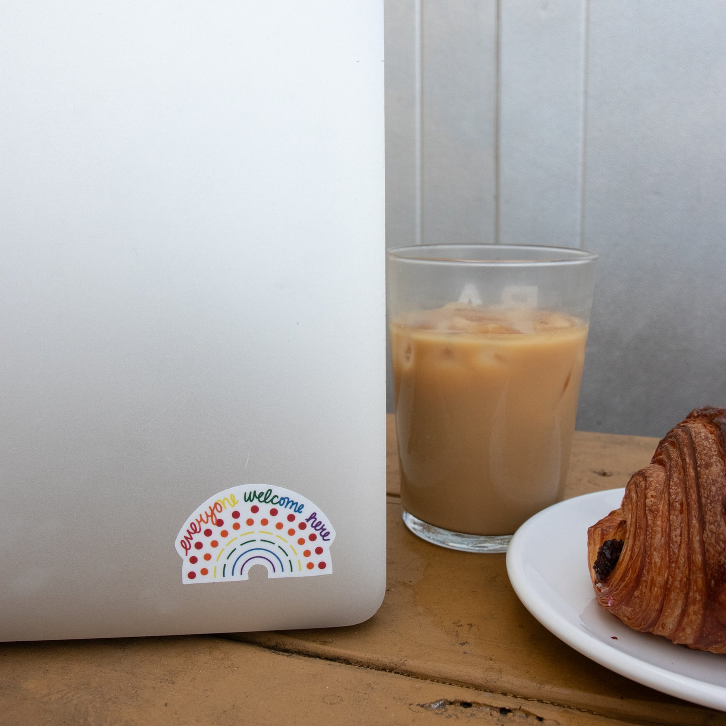 Laptop with a multicolored pride sticker with the phrase everyone welcome here amidst a coffee and croissant