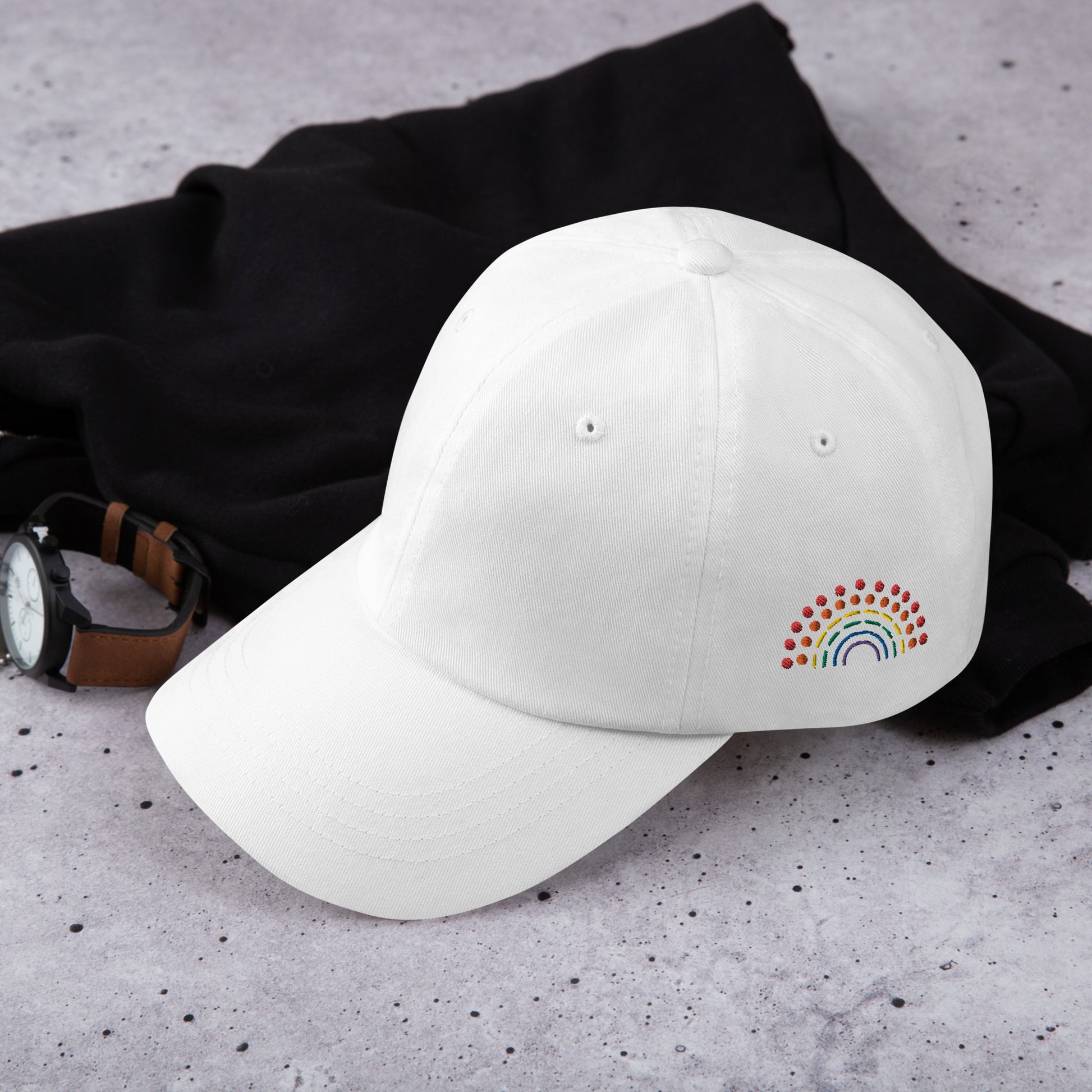 white baseball dad hat with multicolored pride rainbow embroidered on the left of the hat amongst other clothing items